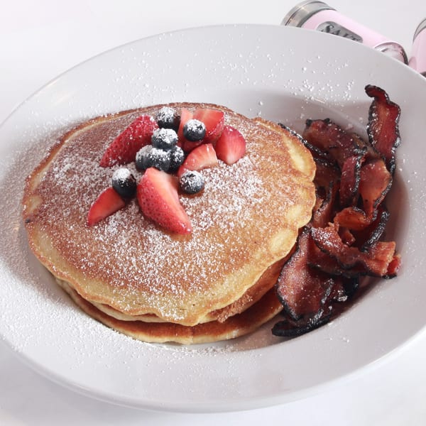 Pancakes with berries and powdered sugar, served with crispy bacon on a white plate.