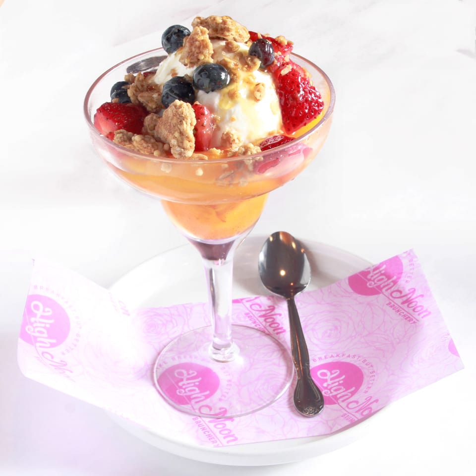 A High Noon Brunchery parfait with yogurt, granola, blueberries, strawberries, and peaches in a glass.