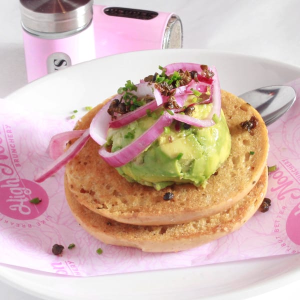 Avocado toast with red onions, chives, and capers on a pink High Noon Brunchery napkin.